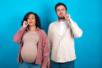 young couple expecting a baby standing against blue background mouth and lips shut as zip with...