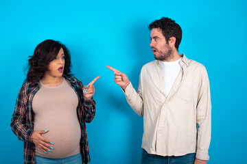Stunned young couple expecting a baby standing against blue background with greatly surprised expression points away on copy space, indicates something