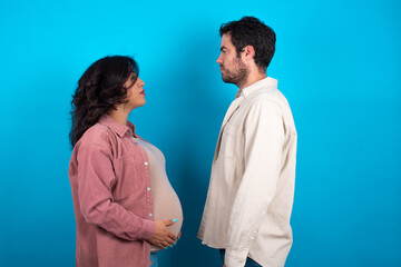 young couple expecting a baby standing against blue background looking to side, relax profile pose with natural face with confident smile.