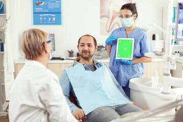 Obraz premium Nurse dentist showing green screen display to stomatology senior doctor while examining tooth pain to man patient sittinh on dental chair. Woman examinating using mock up chroma key tablet