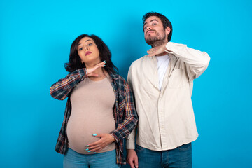 young couple expecting a baby standing against blue background cutting throat with hand as knife,...