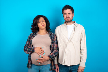 young couple expecting a baby standing against blue background pointing down with fingers showing advertisement, surprised face and open mouth