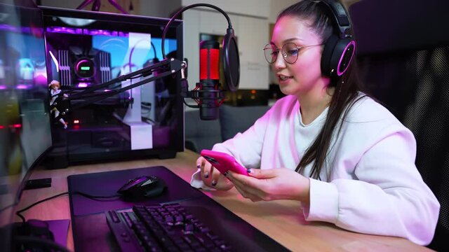 An attractive girl gamer wearing eyeglasses and headphones is streaming online videogames on professional powerful computer while using her smartphone sitting at home