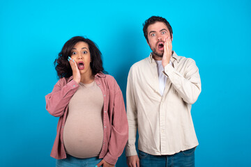 Shocked young couple expecting a baby standing against blue background looks with great surprisment being very stunned, astonished with unexpected news, Facial expressions concept.