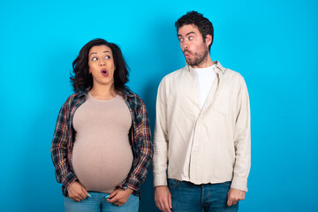 Fototapeta premium Shocked young couple expecting a baby standing against blue background look empty space with open mouth screaming: Oh My God! I can't believe this.