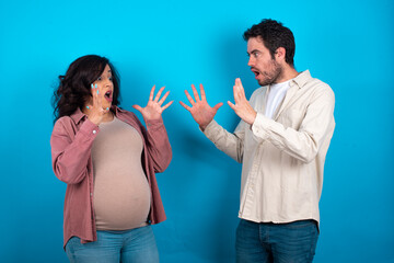 young couple expecting a baby standing against blue background shouts loud, keeps eyes opened and hands tense.