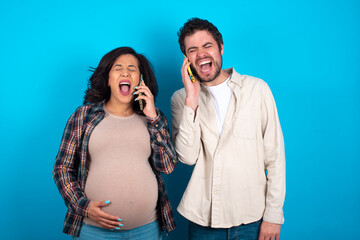 Overemotive happy young couple expecting a baby standing against blue background laughs out positively hears funny story from friend during telephone conversation