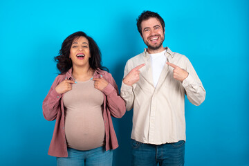 young couple expecting a baby standing against blue background points at his body, being in good mood after going shopping and making successful purchases