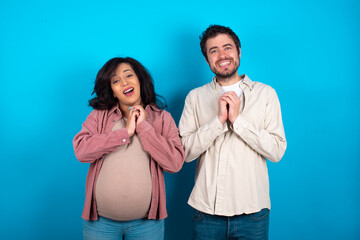 Positive young couple expecting a baby standing against blue background smiles happily, glad to receive pleasant news from interlocutor, keeps hands together. People emotions concept.