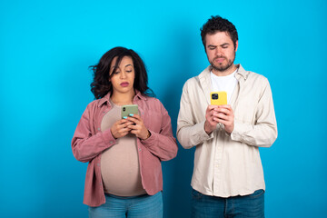 Excited young couple expecting a baby standing against blue background  winking and eye hold smart phone use read social network news