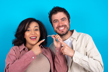 young couple expecting a baby standing against blue background holding an invisible aligner and pointing at it. Dental healthcare and confidence concept.