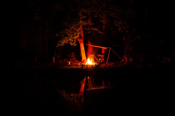 Un feu de bois au bord d´un étang par une nuit sombre. Les flammes éclairent les branches des arbres et le tout se reflète dans l´étang, créant un effet de miroir.