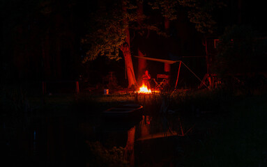 Un feu de camp la nuit au bord d´un étang sous les arbres, une bâche protège une personne assise devant le feu. Les arbres se reflètent dans l´étang.