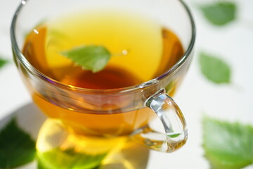 Tea in a glass cup with a birch leaf. Green leaves on a white table around a mug with a drink