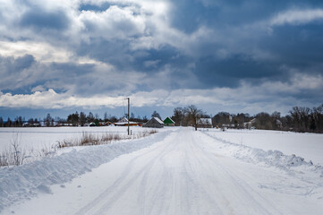 Rural road leading through the fields to the small village. Snowy road with a dramatic sky in the winter season.