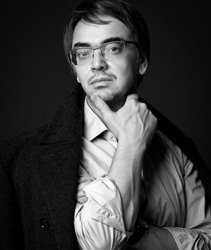 Black and white portrait of thoughtful wise adult man literary critic, poet, writer in glasses, white shirt and teed jacket on shoulders standing holding hand at chin, thinking over black background
