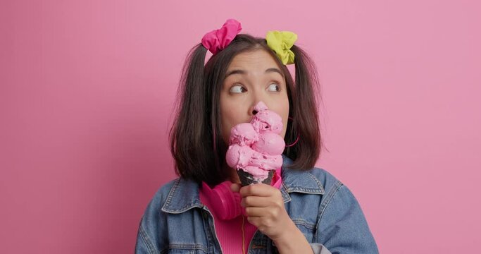 Carefree millennial Asian girl with two pony tails licks delicious ice cream has dirty face dressed in fashionable clothes enjoys spare time or day off poses against pink background. Tasty dessert