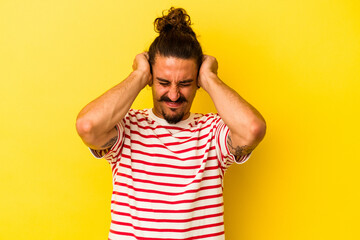 Young caucasian man with long hair isolated on yellow background covering ears with hands.