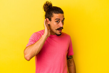 Young caucasian man with long hair isolated on yellow background trying to listening a gossip.