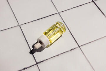 Dropper glass cosmetic bottle with yellow oil or serum for facial spa treatment on a white square tile  bathroom countertop background. Eco-friendly beauty routine.