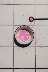 A white bowl and a black teaspoon filled with pink hearts glitter sequins on a white square tile counter background. Celebrating birthday decor. Creative flat lay with copy space.