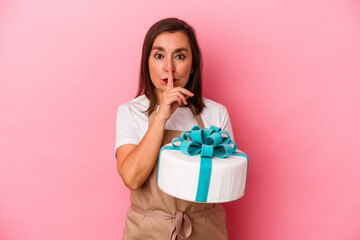 Middle aged pastry chef woman holding a cake isolated on blue background keeping a secret or asking for silence.