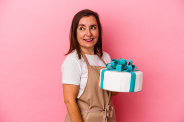 Middle aged pastry chef woman holding a cake isolated on blue background looks aside smiling, cheerful and pleasant.