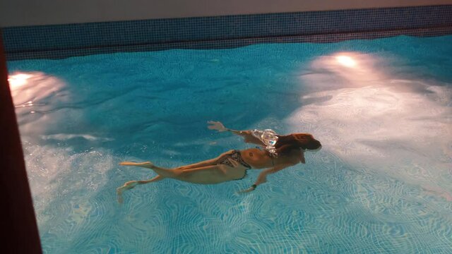 Slow motion shot of a young woman swimming inside of a pool during night time by herself, dark surroundings and pool lights.