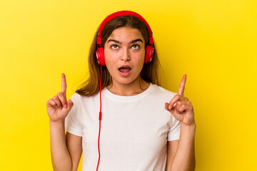 Young caucasian woman listening to music isolated on yellow background pointing upside with opened mouth.