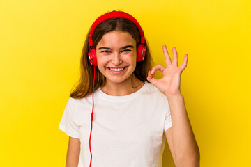 Young caucasian woman listening to music isolated on yellow background cheerful and confident showing ok gesture.