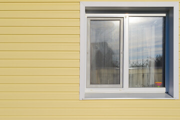 The wall of the house is covered with yellow vinyl siding and a plastic white window