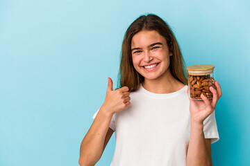 Young caucasian woman holding an almond jar isolated on blue background smiling and raising thumb up