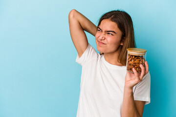 Young caucasian woman holding an almond jar isolated on blue background touching back of head, thinking and making a choice.