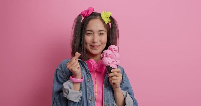 Attractive Asian woman with two pony tails licks appetizing ice cream makes korean like sign wears fashionable denim jacker headphones around neck isolated over pink background. Summer holidays