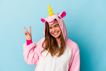 Young caucasian woman wearing a unicorn pajama isolated on blue background joyful and carefree showing a peace symbol with fingers.