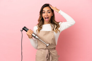 Young woman using hand blender over isolated pink background with surprise expression