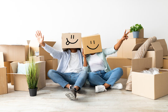Moving house concept. Playful couple with cardboard boxes with smiley faces on head, sitting on floor and having fun
