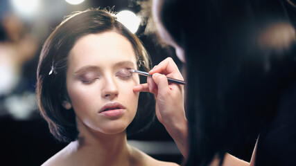 makeup artist applying eye shadow with cosmetic brush on young pretty model with closed eyes