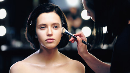 makeup artist applying makeup foundation with cosmetic brush on face of brunette young model