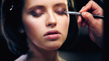makeup artist applying eye shadow with cosmetic brush on eyelids of young woman