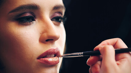 Obraz premium makeup artist applying lip gloss on lips of model isolated on black