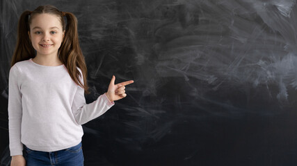 a Caucasian girl, a schoolgirl, is standing against the background of an empty chalk board. the index finger points to an empty space for text. c