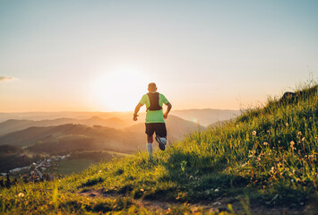 Active mountain trail runner dressed bright t-shirt with backpack running endurance marathon race by picturesque hills at sunset time back view photo. Sporty active people backlight concept image.