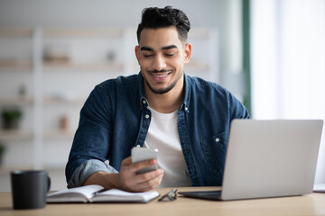 Cheerful arab guy using smartphone while sitting at workdesk