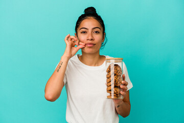 Young latin woman holding a cookies jar isolated on blue background with fingers on lips keeping a...