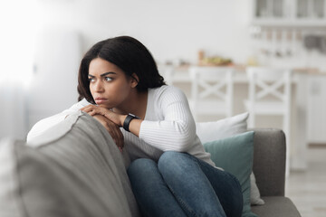 Lonely depressed african american woman sitting on couch with sad face expression and looking away, free space