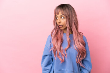 Fototapeta premium Young mixed race woman with pink hair isolated on pink background doing surprise gesture while looking to the side