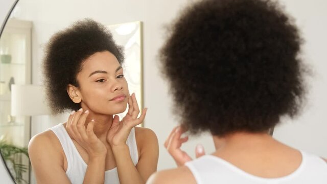 Young African American Black Woman Looking Touching Face In The Mirror Examining Skin Condition And Enjoy Her Healthy Skin. Beauty Product Concept.