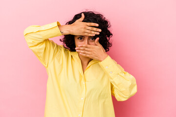 Young curly latin woman isolated on pink background blink at the camera through fingers, embarrassed covering face.