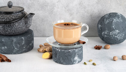 Masala tea chai in a glass cup with traditional teapot on podium or pedestal. Traditional Indian drink beverage - spicy black tea with spices cinnamon, ginger, nutmeg, cardamom, cloves and milk.